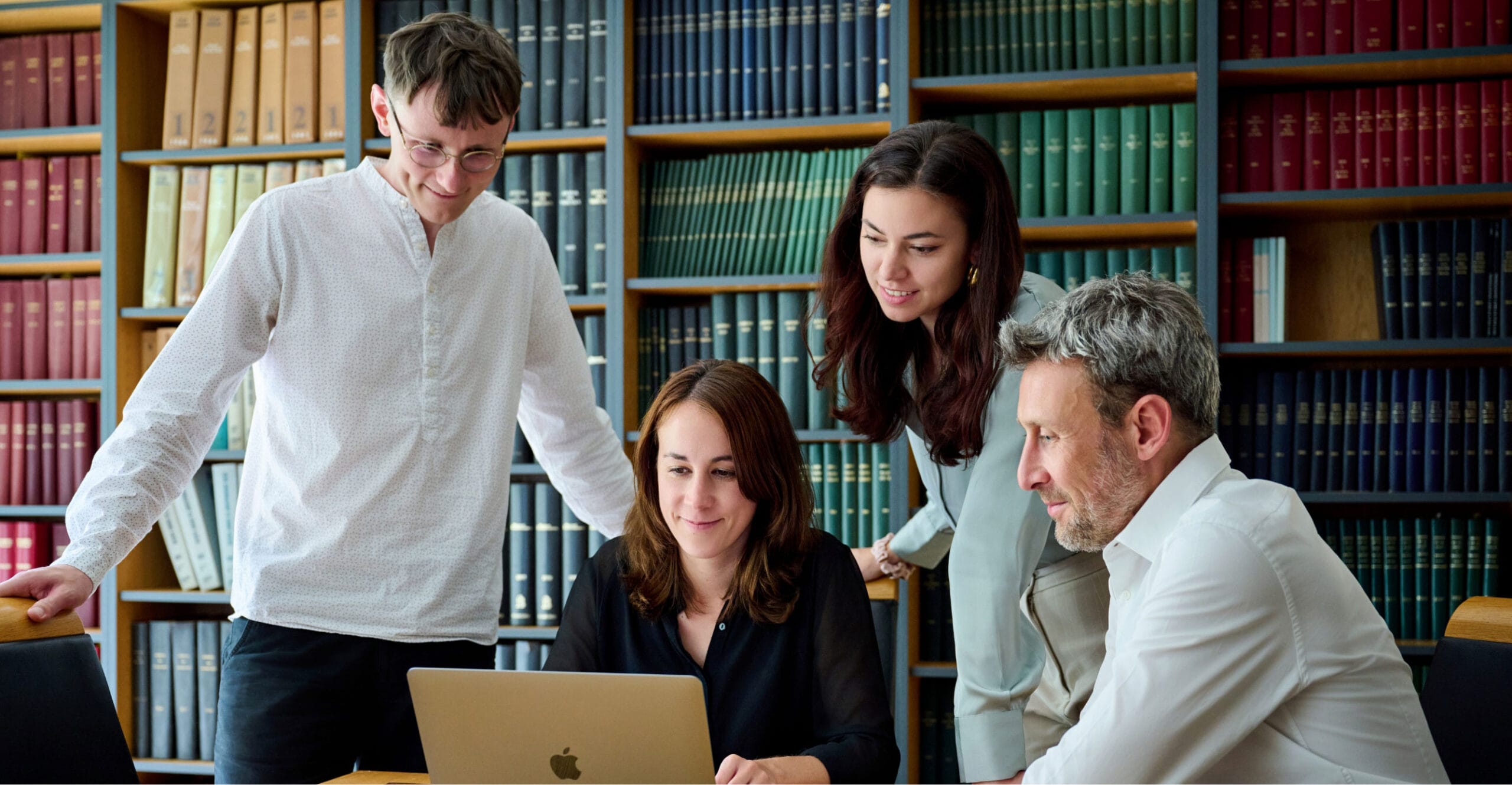 Quatre personnes sont réunies autour d'un ordinateur portable dans une bibliothèque aux étagères remplies de livres. Elles semblent toutes concentrées sur l'écran, absorbées par leur travail ou leur discussion.