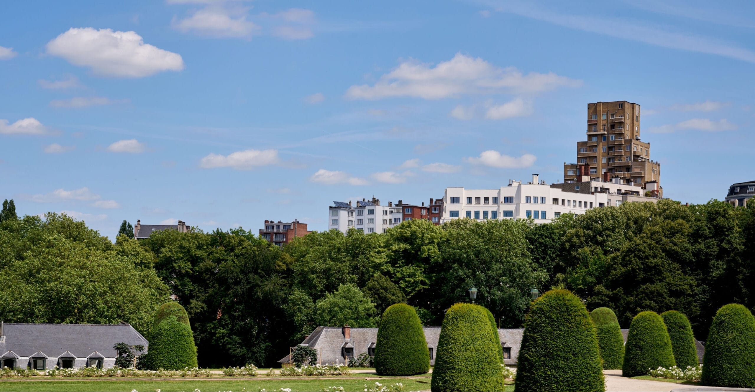 Un parc verdoyant et luxuriant, avec des arbres soigneusement taillés au premier plan. Derrière le parc, plusieurs immeubles bas et un gratte-ciel moderne et original se dressent sous un ciel bleu parsemé de nuages.