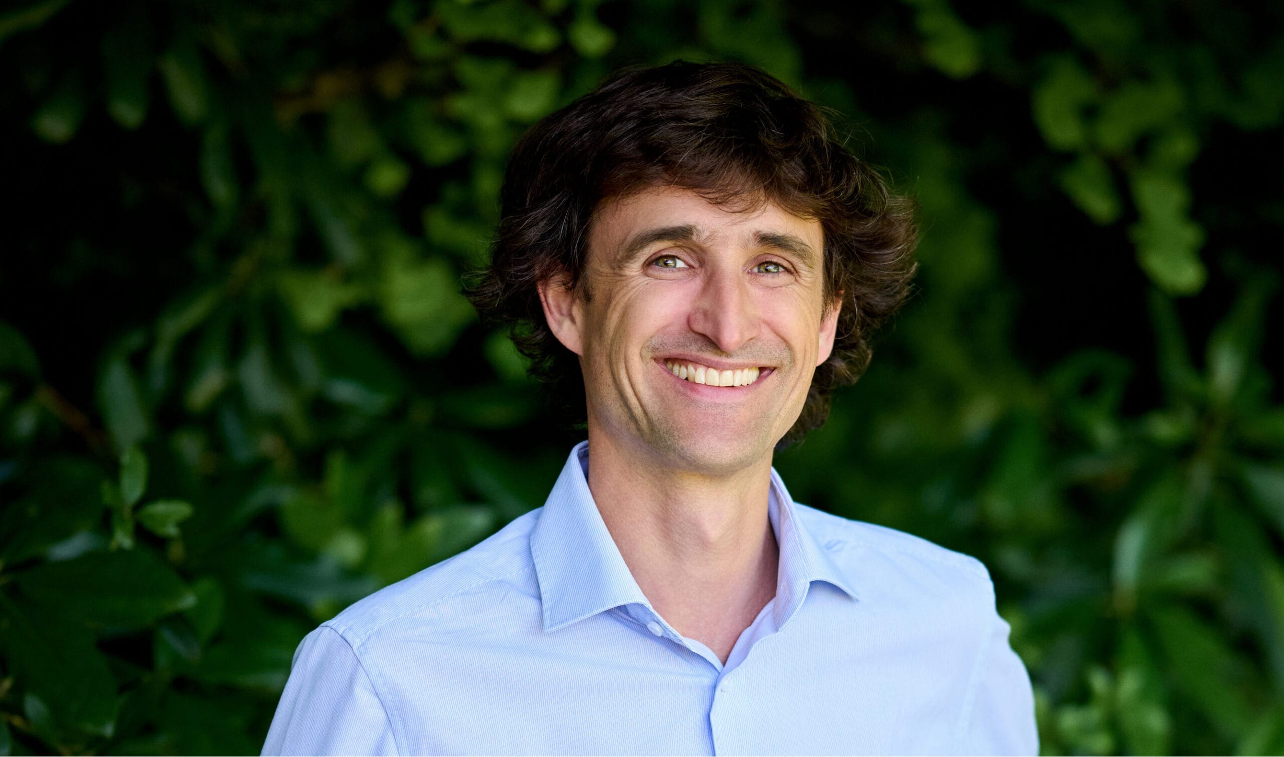 Un homme aux cheveux bruns ondulés, vêtu d'une chemise bleu clair, arbore un large sourire. Il se tient devant un arrière-plan verdoyant et luxuriant.