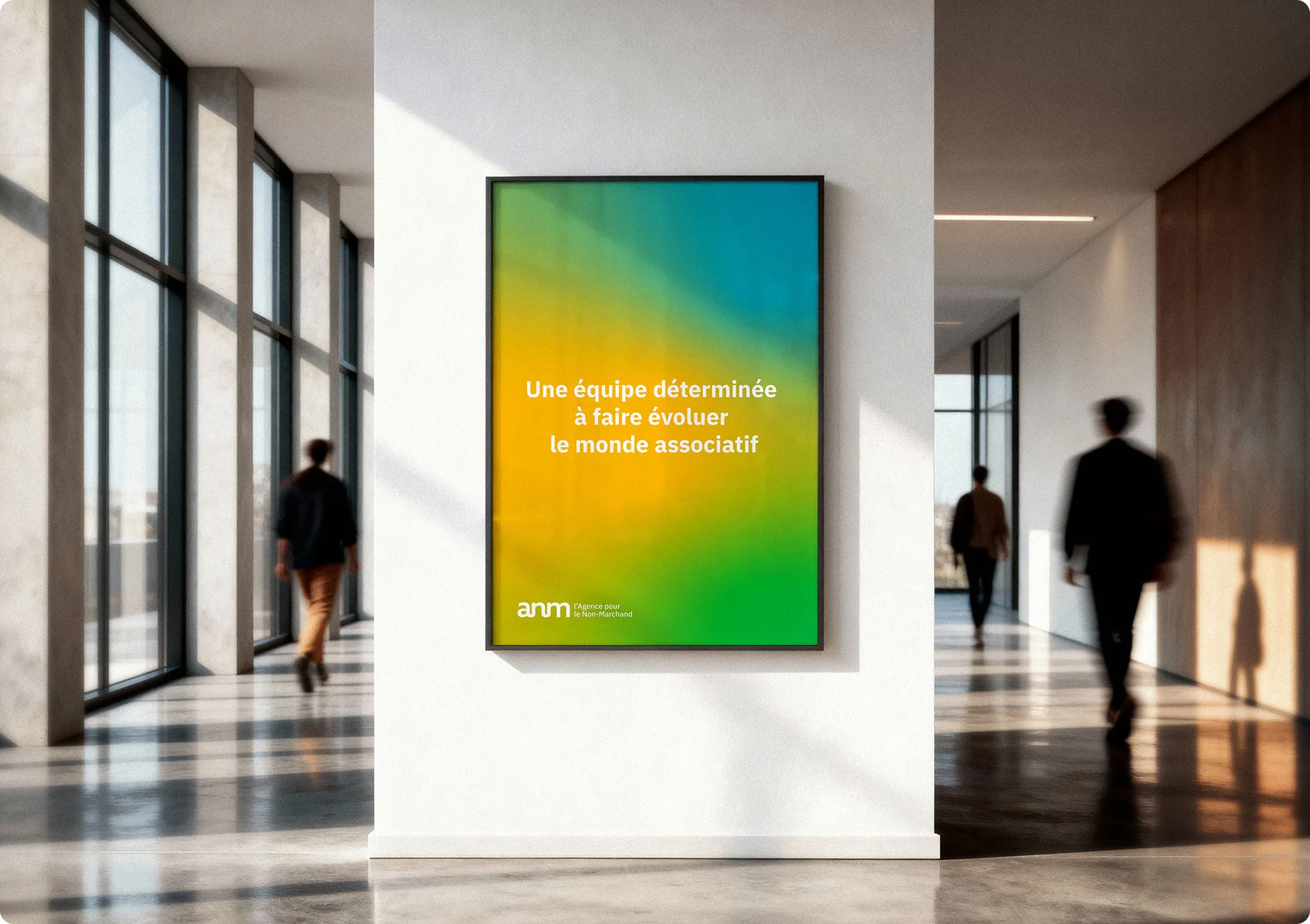 Une affiche encadrée avec un dégradé de vert et de jaune est accrochée au mur blanc d'un couloir moderne. On peut y lire en français : Une équipe déterminée à faire évoluer le monde associatif. Des gens passent à l'arrière-plan.