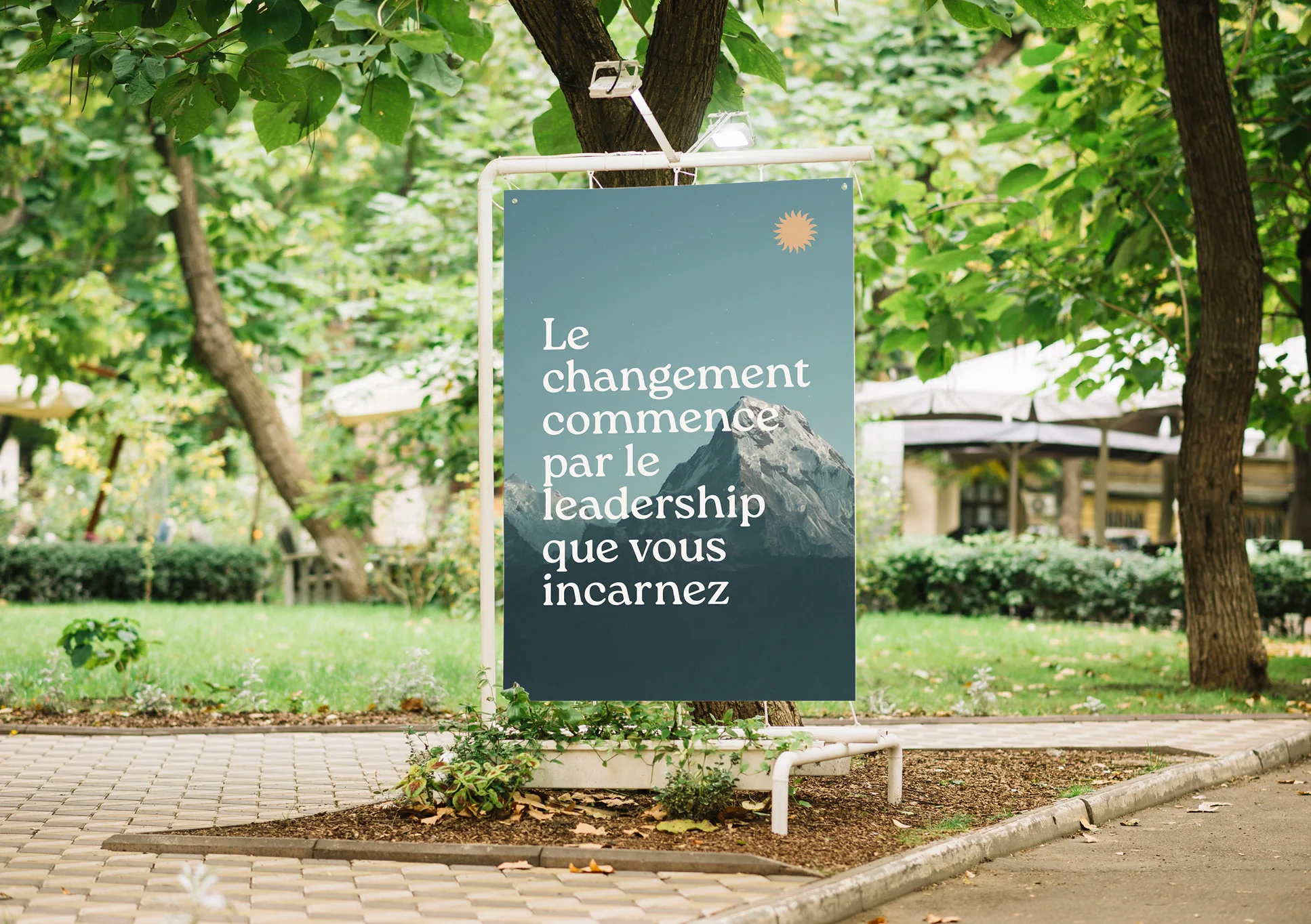 Dans un parc verdoyant, un panneau indique "Le changement commence par le leadership que vous incarnez" en français, avec une petite icône de soleil et l'image d'une montagne en arrière-plan.